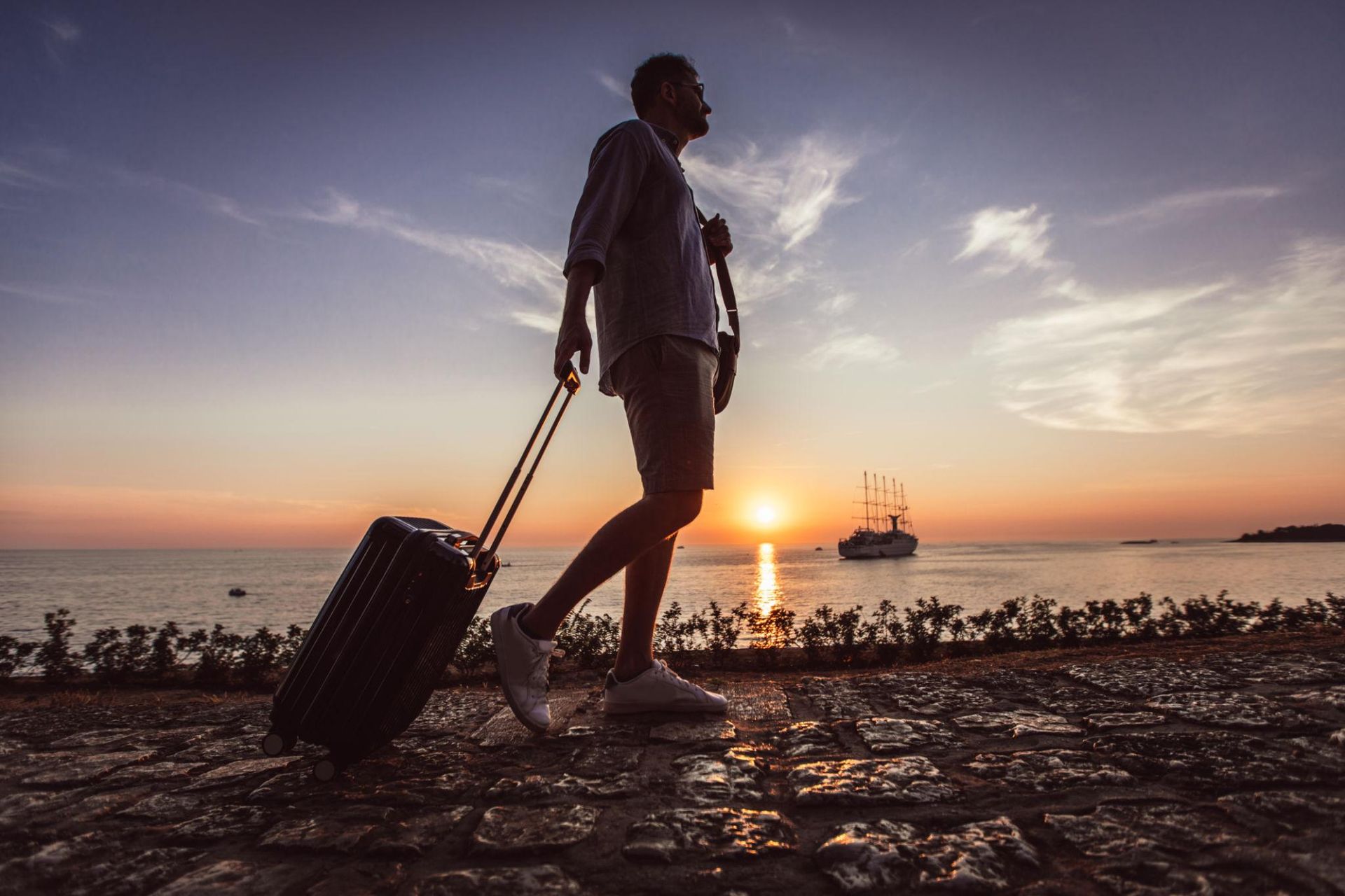 A traveler pulling a suitcase along a seaside promenade at sunset, with a tall ship on the water
